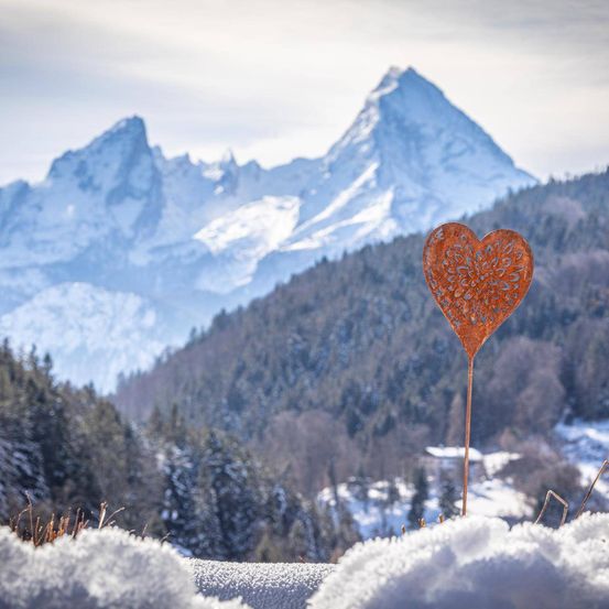 Ein herzförmiges Metalldekoration steht auf einem verschneiten Hügel, mit einer verschneiten Berglandschaft dahinter.