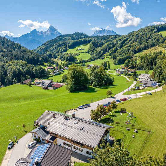 Ein Luftbild eines malerischen Alpendorfs mit Häusern, einem weiten grünen Feld und majestätischen Bergen im Hintergrund unter einem klaren Himmel.