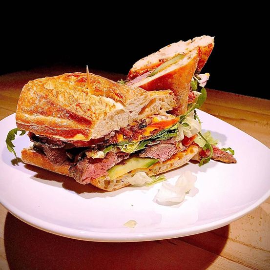 Ein Sandwich mit Fleisch, Käse, Gurke, Salat und Brot liegt auf einem weißen Teller auf einem Holztisch.