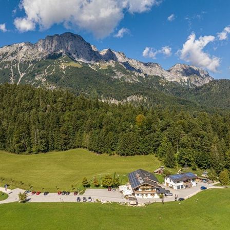 Luftaufnahme eines grünen Tals mit Bergen, einem Haus mit Solarpanelen und einem Parkplatz mit mehreren Autos.