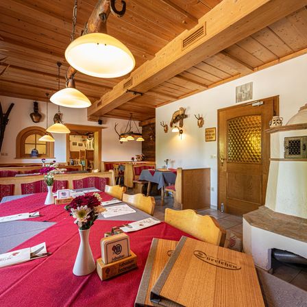 Das Innere eines Essbereichs mit hölzerner Decke, Wänden und Tischen. Es gibt rote Tischdecken, weiße Vasen mit Blumen und Menüs auf den Tischen. Holzbänke und Stühle sind aufgestellt. Eine Tür und ein Kamin sind sichtbar.