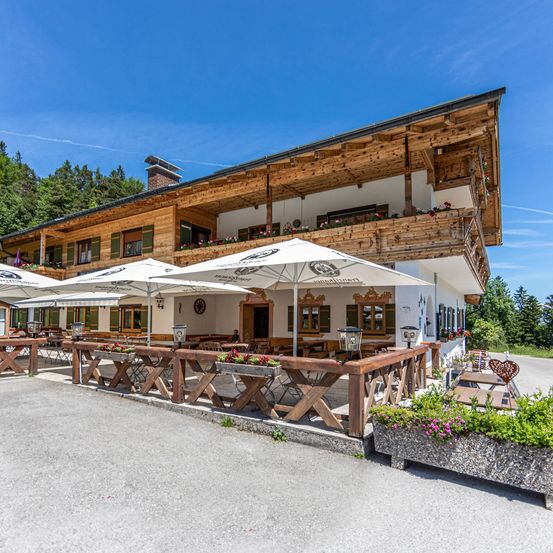 Ein hölzernes Chalet-Stilgebäude mit Balkonen und Fenstern. Außensitzplätze mit weißen Sonnenschirmen. Holzzäune und Tische. Bäume im Hintergrund.