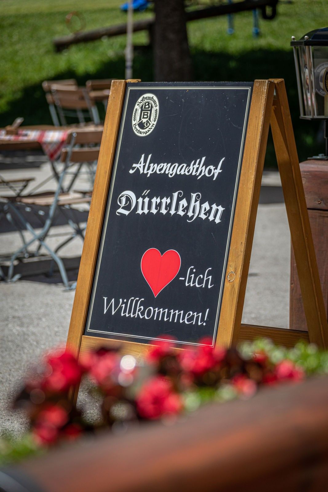 Ein schwarzes Tafelschild mit dem Text 'Alpengasthof Durrleben' und einem roten Herzen. Es steht 'Herzlich willkommen!' Dahinter sind Tische und Stühle.