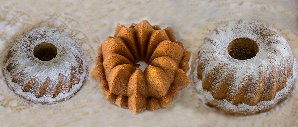 Drei Bundt-Kuchen sind auf einem weißen Tuch ausgestellt. Der mittlere Kuchen hat ein Spiraldesign, während die anderen rund sind und mit Puderzucker bestreut sind.