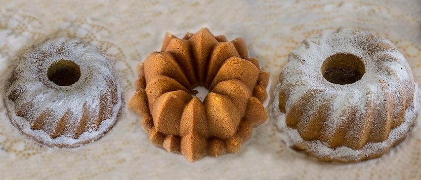 Drei Bundt-Kuchen sind auf einem weißen Tuch ausgestellt. Der mittlere Kuchen hat ein Spiraldesign, während die anderen rund sind und mit Puderzucker bestreut sind.