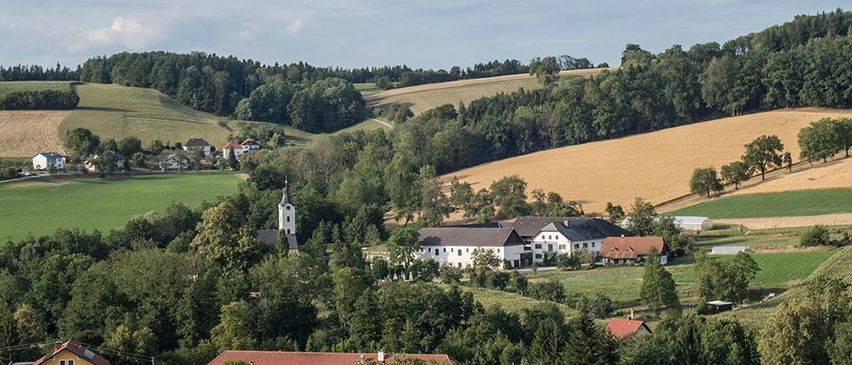 Luftaufnahme einer ländlichen Landschaft mit einer weißen Kirche, einem großen weißen Gebäude und mehreren Häusern, umgeben von Bäumen und Feldern.