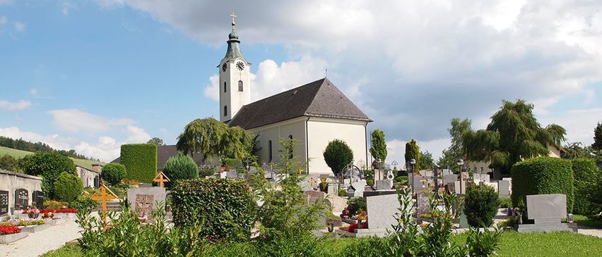 Eine Kirche mit Turm und Uhr steht auf einem Friedhof, umgeben von Bäumen und Büschen.