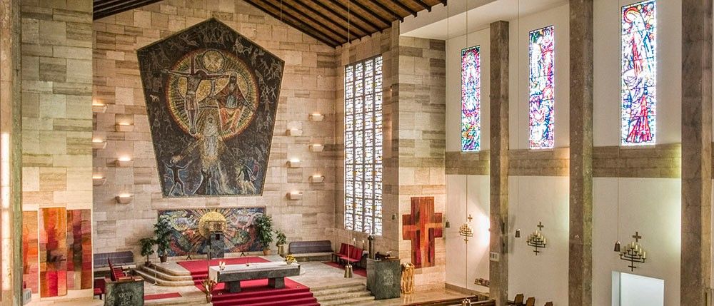 Ein modernes Kircheninneres mit einer großen Mosaikwand, Buntglasfenstern und einem roten Teppich, der zum Altar führt.