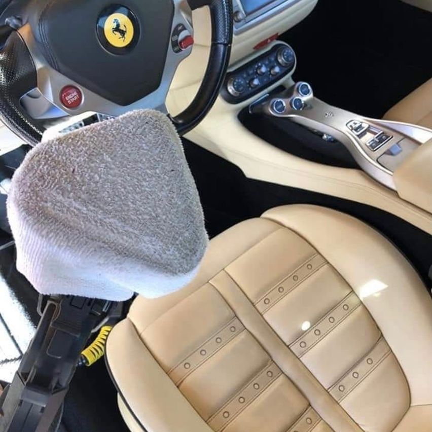 Das Innere eines Ferrari-Sportwagens mit einem beigefarbenen Lederfahrersitz mit sichtbaren Nähten und einem Lenkrad mit Ferrari-Logo. Ein graues Handtuch liegt auf dem Lenkrad.