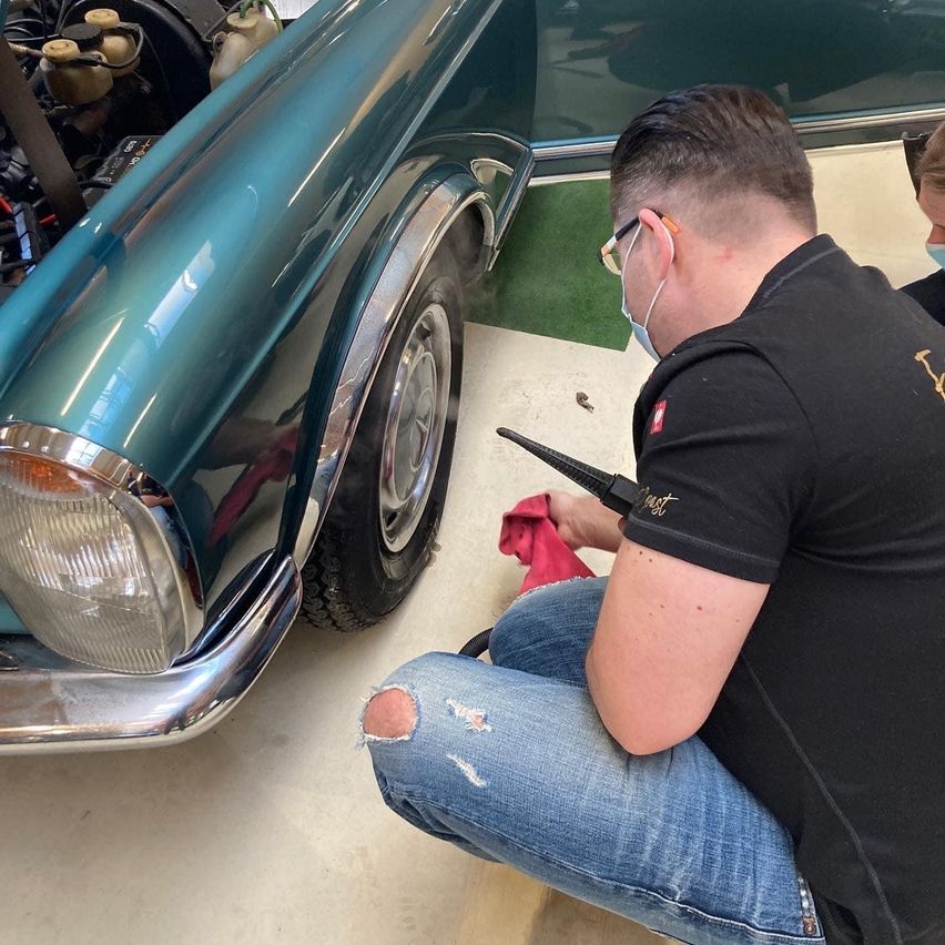 Ein Mann arbeitet an einem Oldtimer und reinigt das Rad mit einem Tuch, während er eine Maske trägt. Der Motor des Autos ist freigelegt.