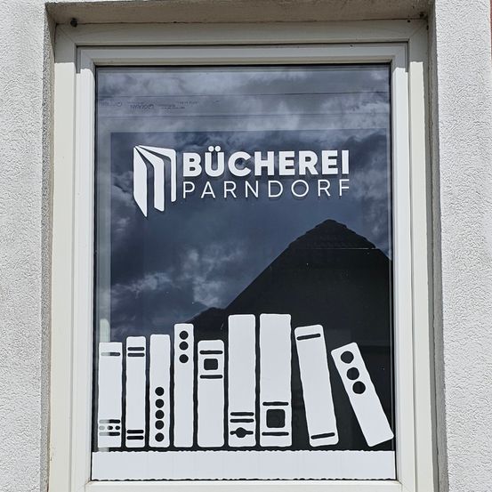 Ein Fenster zeigt ein Logo für Bucherei Parndorf mit einer Reihe von Büchern darunter. Der Hintergrund zeigt einen bewölkten Himmel und einen Berg.