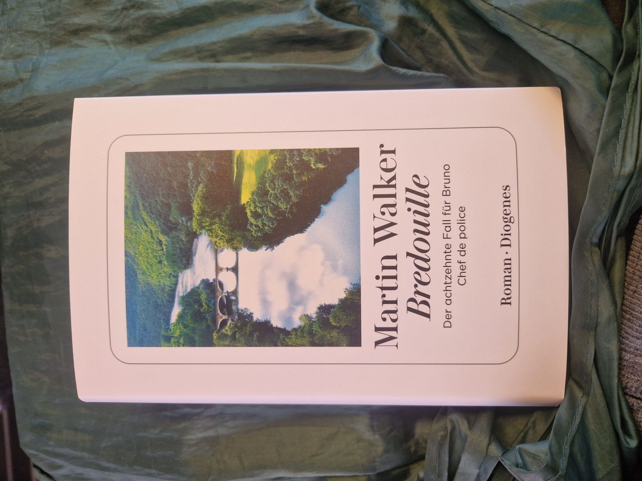 Ein Buchcover mit einer Landschaftsaufnahme, die einen Fluss, eine Brücke und einen bewölkten Himmel zeigt. Der Titel lautet 'Martin Walker Bredoullie' und 'Der achtzehnte Fall für Bruno' steht darunter. Der Name des Autors 'Roman Diogenes' befindet sich unten rechts.