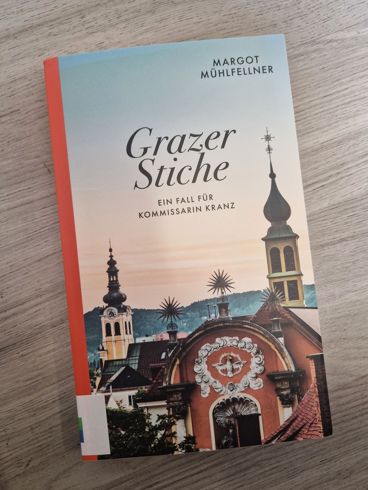 Buchcover mit dem Titel 'Grazer Stiche' und dem Untertitel 'Ein Fall für Kommissarin Kranz'. Das Cover zeigt eine Stadtansicht mit einer Kirche und einem Glockenturm.