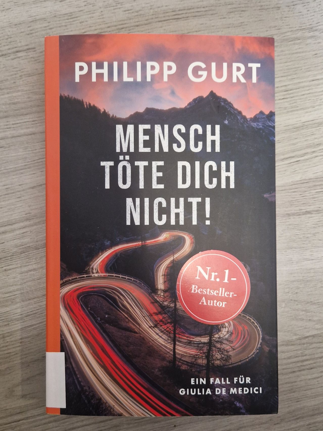 Buchcover mit dem Titel 'Mensch Töte Dich Nicht!' von Philipp Gurt. Es zeigt eine kurvenreiche Straße mit Scheinwerfern und einer Bergkette im Hintergrund. Das Cover hat einen roten Aufkleber, der 'Nr. 1-Bestseller-Autor' lautet.