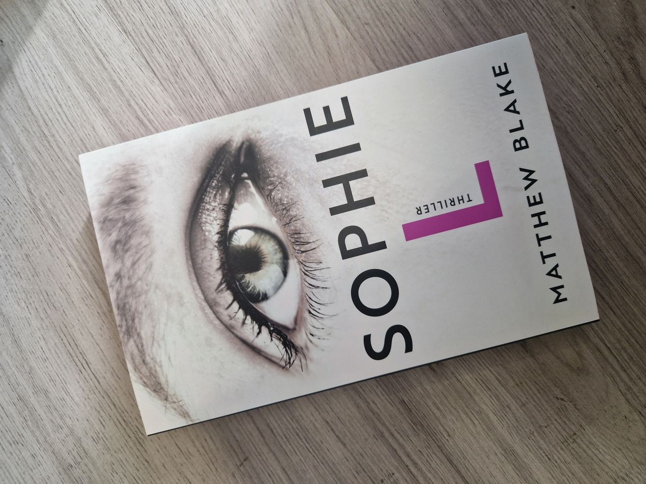 Ein Buchcover mit einer Nahaufnahme eines menschlichen Auges mit dem Titel 'Sophie' und dem Namen des Autors 'Matthew Blake'. Das Auge ist in Schwarz-Weiß mit einem rosafarbenen Highlight auf den Wimpern.