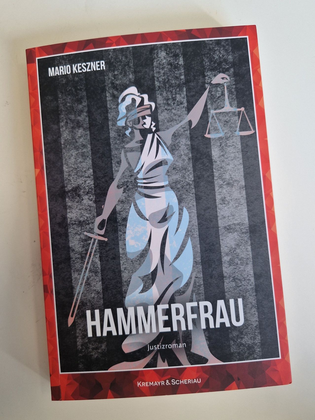 Ein Poster mit einer verblindeten Frau, die einen Schwert und eine Waage hält, mit dem Titel 'Hammerfrau' und dem Namen des Autors 'Mario Keszner' darüber. Das Poster hat einen schwarz-weiß gestreiften Hintergrund.
