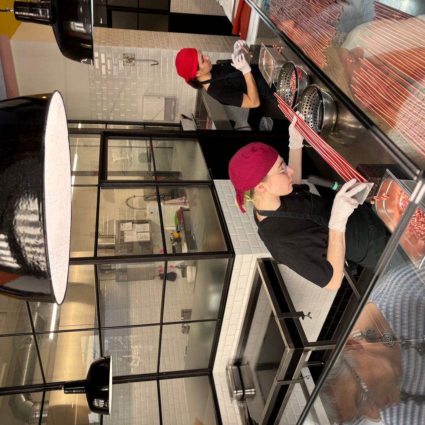 Eine Küche mit zwei arbeitenden Personen, einer mit rotem Hut, der einen Stock hält, und einer anderen mit schwarzem Shirt und Handschuhen. Eine Person reflektiert sich auf dem Glas.