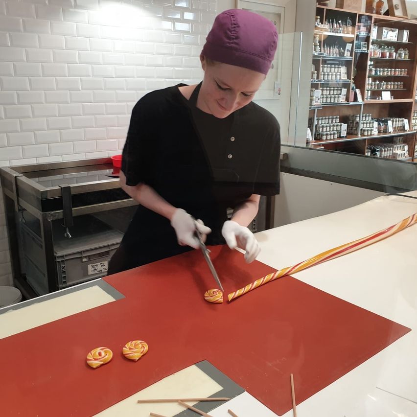 Eine Frau mit Kochmütze und Handschuhen schneidet einen Zuckerrohrstab auf einer roten Oberfläche. Sie befindet sich in einem Raum mit weiß gefliester Wand und einem Regal mit vielen Flaschen.