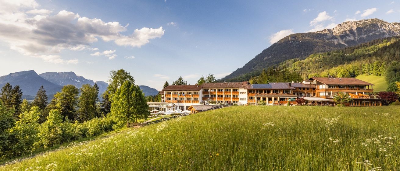 Ein Hotel thront auf einem grasbewachsenen Hügel mit Bergen und Bäumen im Hintergrund unter einem klaren blauen Himmel.