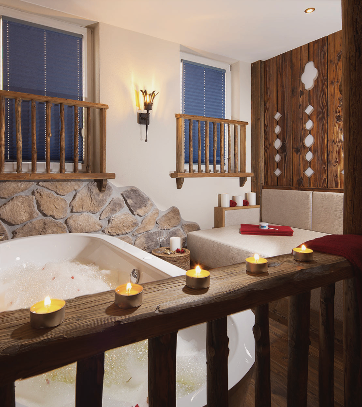 Ein holzverkleideter Raum mit einer großen Badewanne gefüllt mit Blasen. Es hat Holzbalkone und Wände. Es gibt Kerzen auf der Balusterbrüstung.