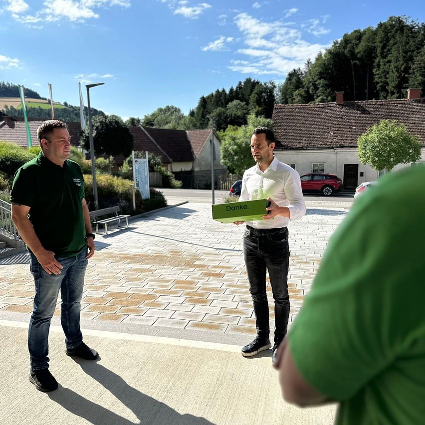 Drei Männer stehen auf einem gepflasterten Gehweg, einer hält eine grüne Box mit der Aufschrift 'Danke'. Im Hintergrund befinden sich Bäume und Häuser.