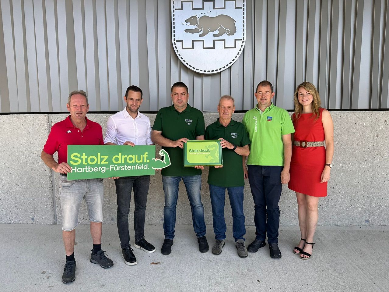 Fünf Erwachsene posieren vor einem Gebäude mit Logo, halten ein grünes Schild mit 'Stolz drauf. Hartberg-Fürstenfeld.' Die beiden Männer an den Enden tragen rote und weiße Hemden.
