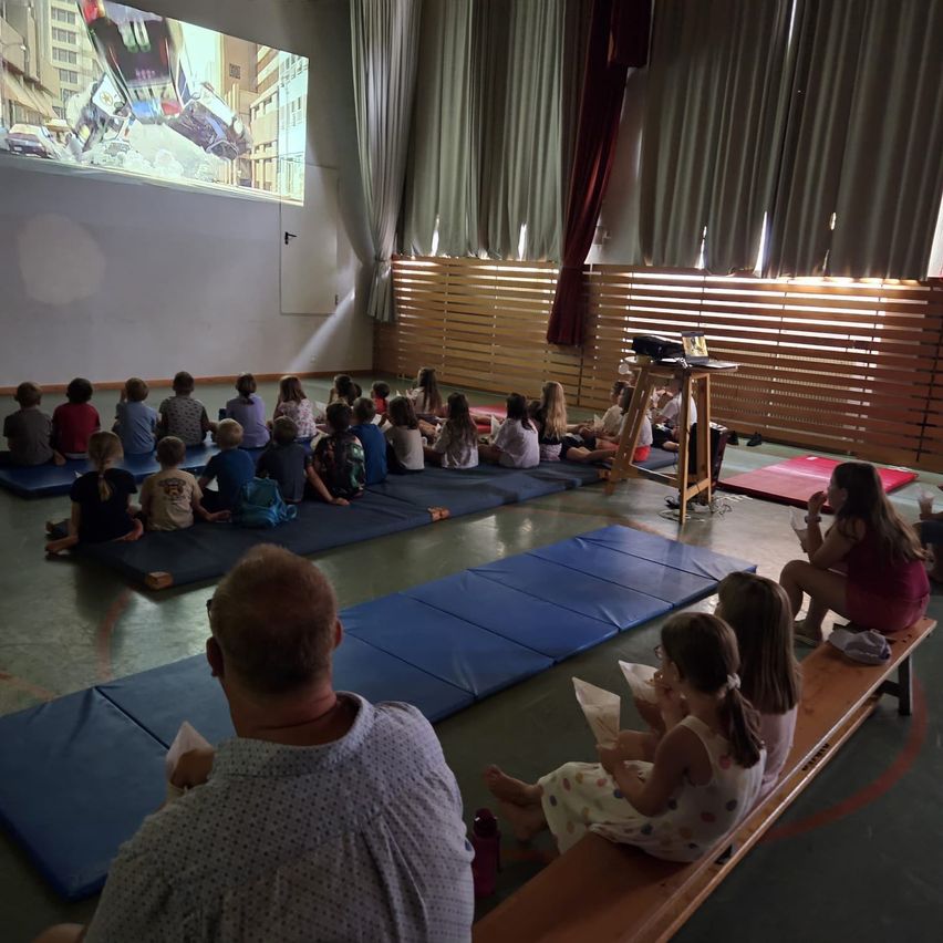 Eine Gruppe von Kindern und Erwachsenen sitzt auf blauen Matten und schaut sich einen Film auf einer großen Leinwand in einem Raum mit Holzwänden und Vorhängen an.