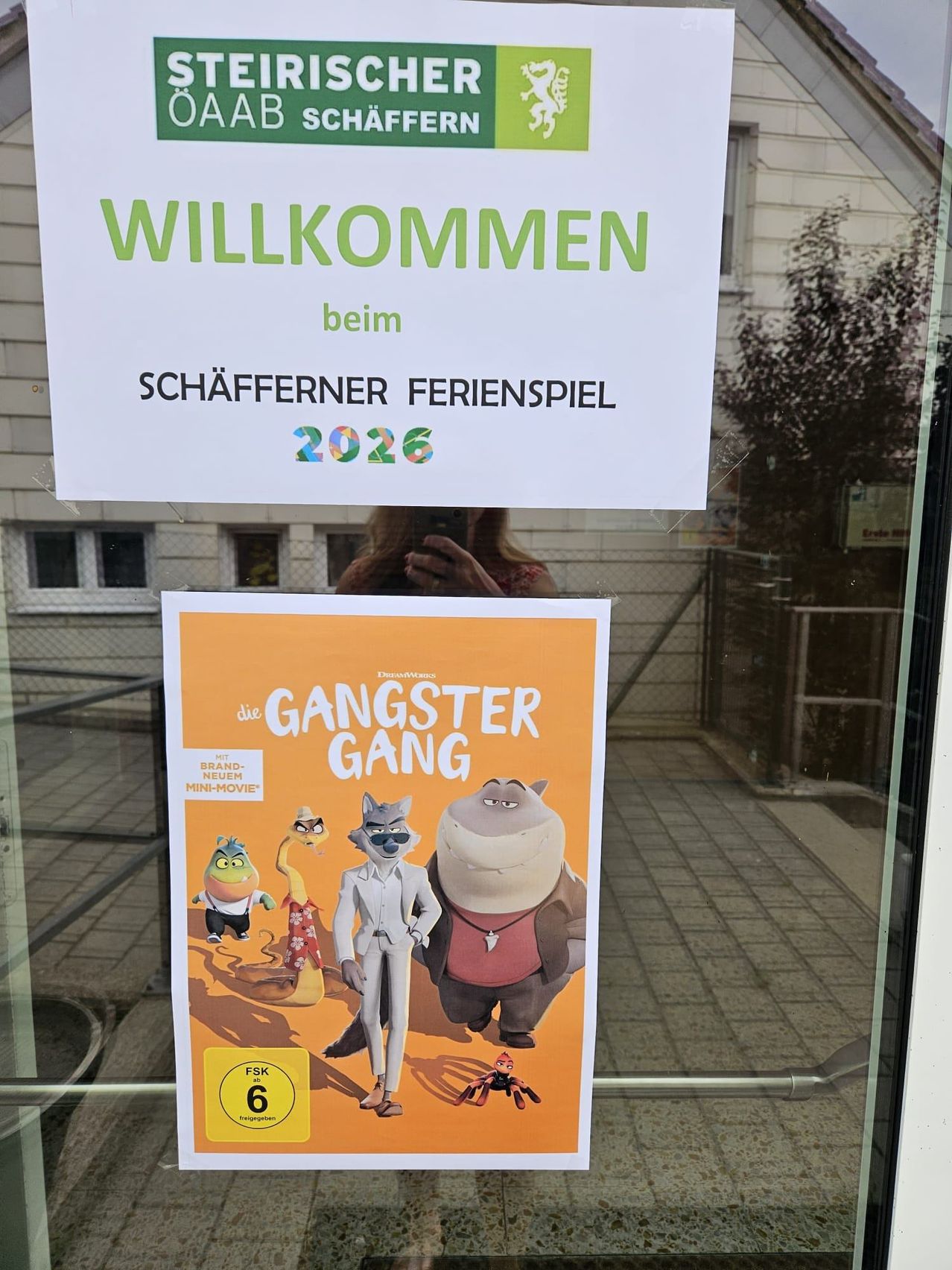 Ein Plakat für den animierten Film 'Die Gangster Gang' ist an einer Glastür angebracht. Darüber steht auf einem weißen Schild 'Willkommen beim Schäfferner Ferienspiel 2026'. Der Spiegel einer Person ist auf der Scheibe zu sehen.