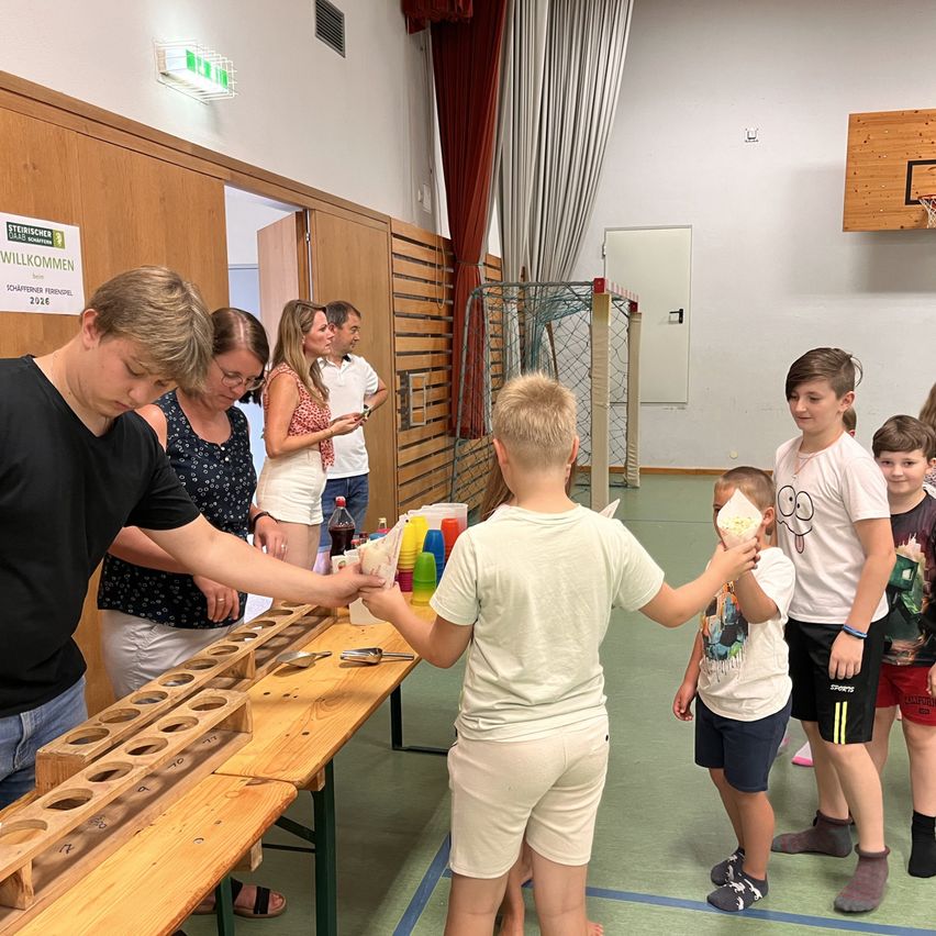 Eine Gruppe von Kindern und Erwachsenen versammelt sich um einen Tisch mit Bechern und einer Spieleinrichtung, möglicherweise an einer Schulveranstaltung teilnehmend.