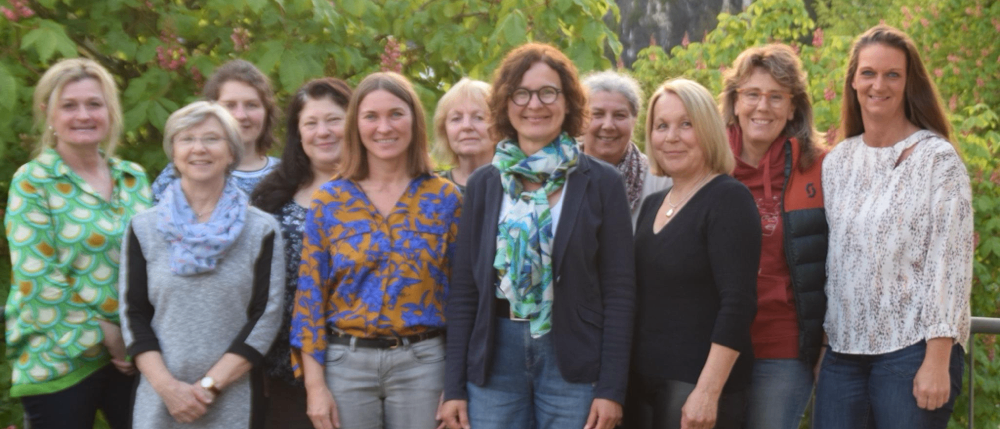 Eine Gruppe von Frauen steht im Freien in der Nähe eines Felsens mit üppiger Vegetation. Sie tragen Freizeitkleidung und scheinen für ein Foto zu posieren.