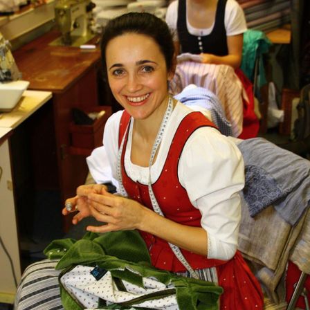 Eine Frau in einem roten und weißen Kleid mit einem Maßband um den Hals lächelt, während sie sitzt und ein grünes Kleidungsstück näht. Hinter ihr näht eine weitere Person, und der Raum ist mit Nähmaschinen und Stoff gefüllt.