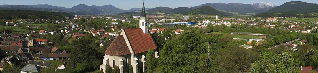 Luftaufnahme einer Stadt mit einer Kirche, umgeben von Bäumen und Bergen. Die Kirche hat ein rotes Dach und einen Turm. Die Stadt ist dicht bebaut mit vielen Häusern und Gebäuden.
