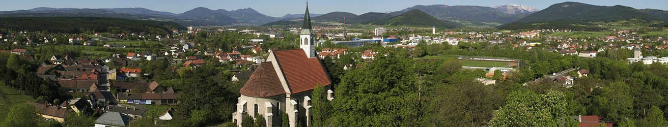 Luftaufnahme einer Stadt mit einer Kirche, umgeben von Bäumen und Bergen. Die Kirche hat ein rotes Dach und einen Turm. Die Stadt ist dicht bebaut mit vielen Häusern und Gebäuden.