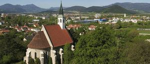 Luftaufnahme einer Stadt mit einer Kirche, umgeben von Bäumen und Bergen. Die Kirche hat ein rotes Dach und einen Turm. Die Stadt ist dicht bebaut mit vielen Häusern und Gebäuden.