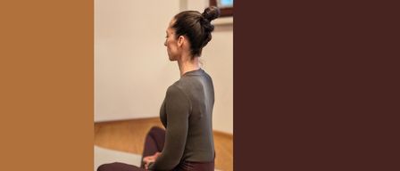 Eine Frau mit Dutt sitzt mit überkreuzten Beinen auf einer Matte in einem Raum und blickt in die Ferne. Sie trägt ein graues Langarmshirt und eine kastanienbraune Hose.