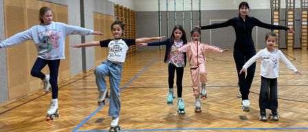 Eine Gruppe junger Mädchen und eine Frau sind auf einem Holzboden rollerskating. Sie tragen Rollschuhe und halten sich an den Händen. Sie alle lächeln und scheinen Spaß zu haben.