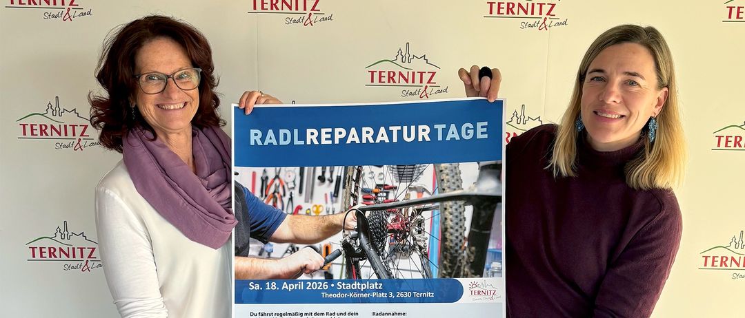 Zwei Frauen stehen vor einer Wand mit TERNITZ-Logos. Eine hält ein Poster mit einem blau-weißen Design, das ein Fahrrad und Werkzeug zeigt. Sie tragen Winterkleidung, und die Frau auf der rechten Seite hat Ohrringe und eine Uhr. Die andere trägt Stiefel.
