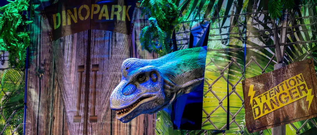 Ein Dinosaurierkopf ragt aus einer Holzwand mit einem Schild 'Dinopark' darüber. Die Szene ist in einem Dschungel mit grünen Pflanzen und Stacheldraht.