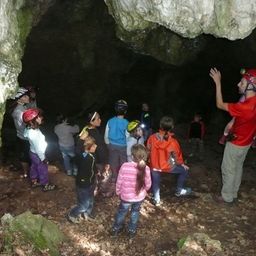 Eine Gruppe von Kindern und Erwachsenen erkundet eine Höhle mit Helmen und Lampen, ein Mann in Rot gestikuliert.