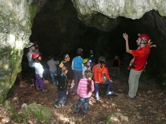 Eine Gruppe von Kindern und Erwachsenen erkundet eine Höhle mit Helmen und Lampen, ein Mann in Rot gestikuliert.