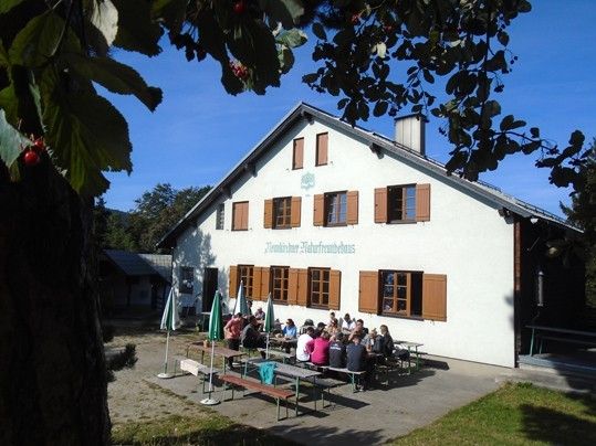 Eine Gruppe von Menschen sitzt an Picknicktischen vor einem weißen Gebäude mit braunen Fensterläden, unter grünen Sonnenschirmen. Das Gebäude hat ein Schild über dem Eingang.