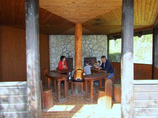 Eine Familie sitzt in einem hölzernen Pavillon mit Ziegelboden und Steinwand. Der Vater sitzt mit zwei Kindern, während die Mutter in der Nähe steht.
