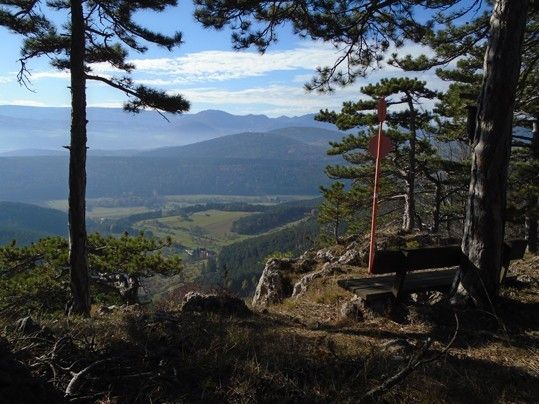 Auf einem felsigen Grat bietet eine Bank einen Blick auf sanfte Hügel, üppige Täler und entfernte Berge unter einem blauen Himmel mit verstreuten Wolken.