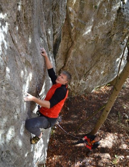 Ein Mann klettert an einer Felswand und trägt eine rot-schwarze Jacke und graue Hose. Eine weitere Person steht unter ihm und hält ein Seil. Sie befinden sich in einem Wald.