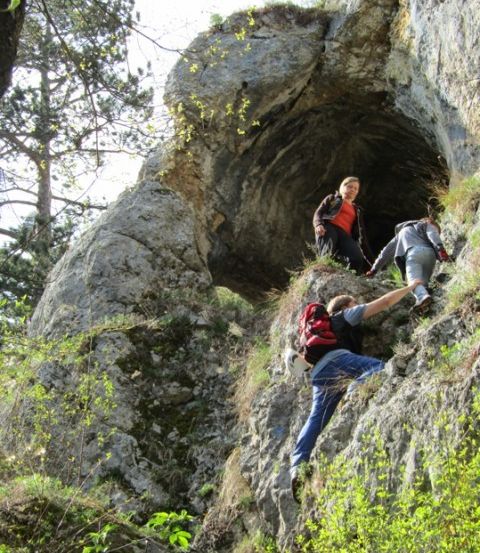 Drei Personen erklimmen einen steilen felsigen Felsen. Die Person auf der linken Seite hilft einer anderen beim Klettern. Die Frau in der Mitte steht am Eingang einer Höhle.