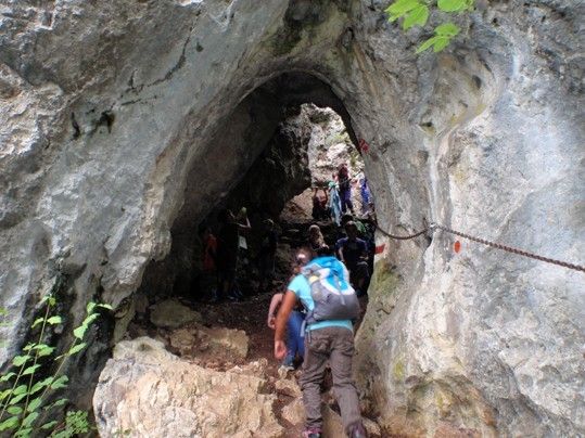 Eine Gruppe von Wanderern bewegt sich durch einen engen Höhleneingang, der durch eine Kette gesichert ist. Sie erklimmen felsige Stufen und tragen Rucksäcke.