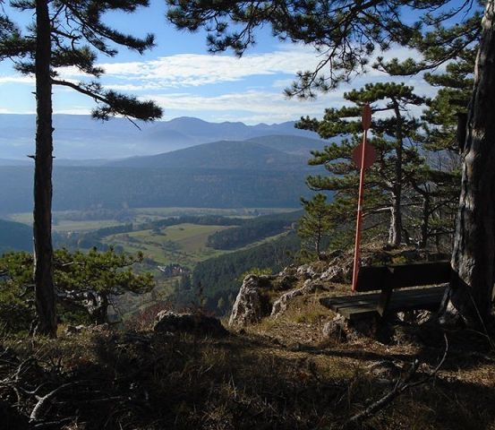 Ein Panoramablick vom Berggipfel zeigt eine Holzbank, ein rotes Schild und ein weites grünes Tal unter einem blauen Himmel mit Wolken.
