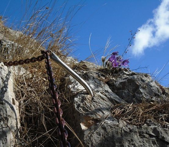 Eine Kette ist an einem Felsvorsprung befestigt, mit violetten Blumen, die in den Felsspalten wachsen. Der Himmel ist blau mit einigen Wolken.