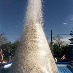 Ein kegelförmiger Wasserbrunnen sprüht Wasser im Freien. In der Nähe stehen einige Leute, einige stehen an der Seite.