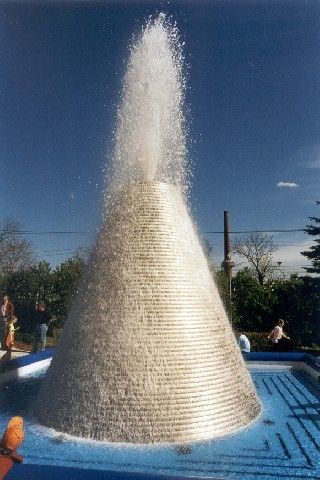Ein kegelförmiger Wasserbrunnen sprüht Wasser im Freien. In der Nähe stehen einige Leute, einige stehen an der Seite.